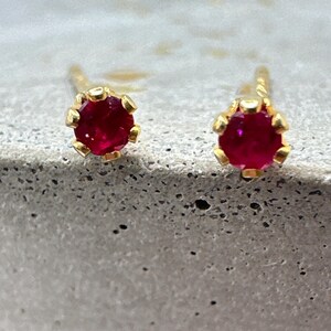 May include: A pair of gold stud earrings, each featuring a vibrant red gemstone. The gemstones are round and set in a gold bezel setting. The earrings are displayed on a textured gray surface.