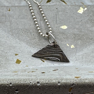 Puede incluir: Un collar de cadena de plata con un colgante triangular hecho de metal estampado. El colgante tiene un esquema de color gris oscuro y plateado.