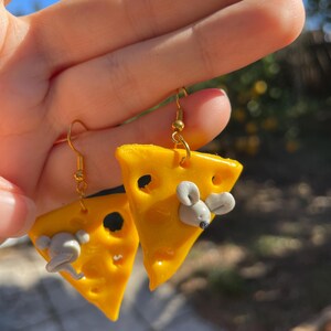 May include: A pair of gold dangle earrings featuring a yellow cheese wedge with a small gray mouse on top. The cheese has holes and is a realistic depiction of a slice of cheese.