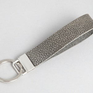 Könnte beinhalten: Graue Leder-Schlüsselanhänger-Handschlaufe mit strukturierter Oberfläche. Der Riemen ist an einem silberfarbenen Metallring und einem rechteckigen Metallstück befestigt.