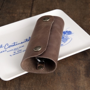 Könnte beinhalten: Ein brauner Leder-Schlüsselanhänger mit Metall-Akzenten. Der Schlüsselanhänger ist aufgerollt und liegt auf einem weißen Keramikteller mit blauem Text, der "hotel Continental Loano Riviera delle Palme Italia 0182 833333 Continental Hotel" lautet.