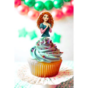 Op de afbeelding: Een cupcake met lichtblauwe glazuur en kleurrijke sprinkles, bekroond met een cartoonafbeelding van een personage met rood krullend haar. De cupcake staat op een witte papieren vorm, met groene en roze ballonnen op de achtergrond.