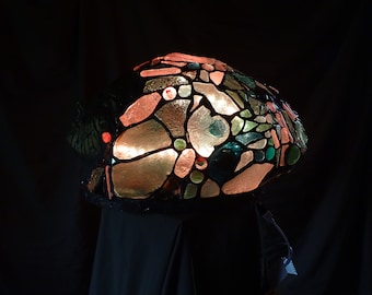 Puede incluir: Una pantalla de lámpara de vidrio coloreado con un diseño de mosaico en tonos de rosa, verde y marrón. La pantalla está iluminada desde el interior, creando un brillo cálido.