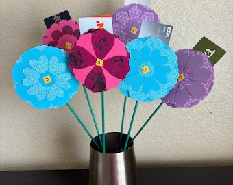 May include: A bouquet of paper flowers in a silver vase. The flowers are blue, purple, pink, and red. The flowers are made of paper and have a 3D effect. The vase is silver and has a round shape.