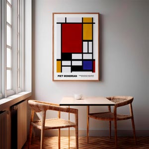 以下が含まれることがあります： ピエト・モンドリアンの幾何学的な抽象画の額装されたプリント。赤、黄、青、黒、白の正方形と長方形の構成が特徴です。プリントは白い壁にかけられ、木製のテーブルと2つの椅子の上にあります。