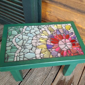 Pode incluir: Um pequeno banco retangular de madeira verde com um tampo de mosaico. O mosaico apresenta um desenho floral com azulejos vermelhos, rosa, amarelos e roxos, e um desenho abstrato em verde claro e branco.