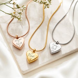 May include: Three heart-shaped pendant necklaces with snake chains in rose gold, yellow gold, and silver tones. Each pendant features custom black script engraving: Mum + Dad x, Nan x, and Love Dad xx, respectively.