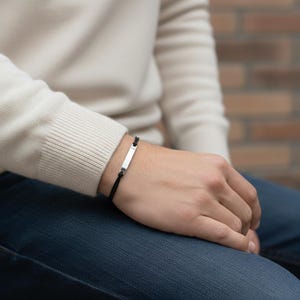 May include: A minimalist bracelet featuring a thin black cord and a small, rectangular silver-toned metal bar centered on the wrist. The cord is secured with simple knots on either side of the metallic plate, creating a clean and understated look.
