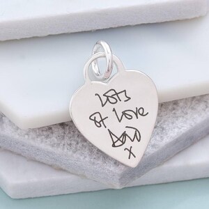 A silver heart-shaped charm with handwritten text on it, resting on a white surface.