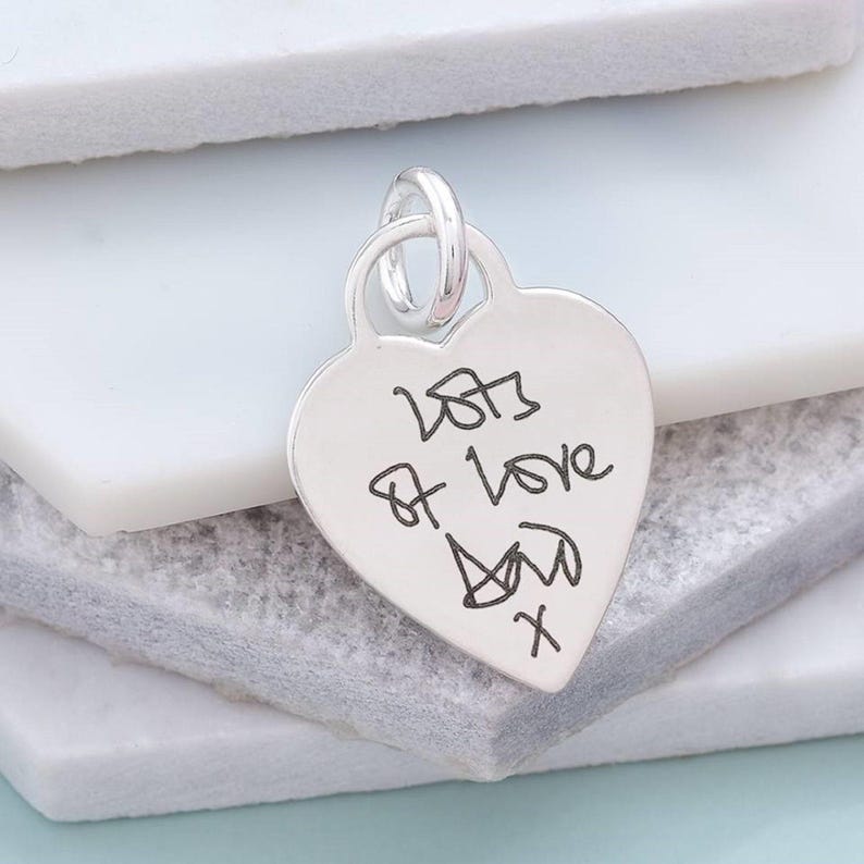 A silver heart-shaped charm with handwritten text on it, resting on a white surface.