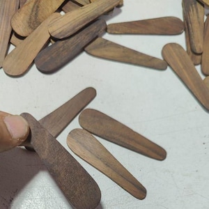 Könnte beinhalten: Eine Sammlung kleiner, holzfarbener Spatel in Tropfenform in verschiedenen Brauntönen. Einige Spatel werden von einer Hand gehalten. Die Spatel sind auf einer weißen Oberfläche angeordnet.