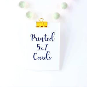 Puede incluir: Una tarjeta blanca con el texto "Printed 5x7 Cards" escrito en cursiva azul. La tarjeta est&aacute; unida a un fondo blanco con una grapa dorada y una cadena de pompones blancos y verdes.