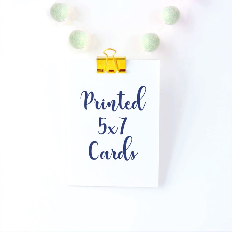 Puede incluir: Una tarjeta blanca con el texto "Printed 5x7 Cards" escrito en cursiva azul. La tarjeta est&aacute; unida a un fondo blanco con una grapa dorada y una cadena de pompones blancos y verdes.