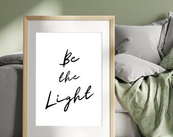 May include: A framed print with a white background and black text that reads "Be the Light". The print is hanging on a wall in a room with a green wall and a couch with pillows.