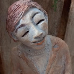 Könnte beinhalten: Keramikskulptur einer sitzenden Figur mit geschlossenen Augen und leichtem Lächeln. Die Figur trägt eine braune Jacke und hat einen hellen Hautton. Die Skulptur ruht auf einem Holzblock.