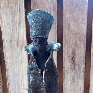May include: A ceramic sculpture of a person wearing a tall, blue hat and a long, dark robe with gold accents. The sculpture has a textured surface and is standing on a wooden base.