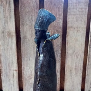 May include: A ceramic sculpture of a person wearing a blue and brown robe with a tall, blue hat. The sculpture has a textured surface and is standing on a woven mat.
