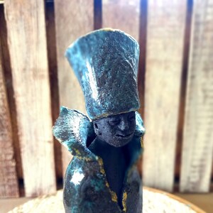 May include: A ceramic figurine of a person wearing a teal and black hat and a teal and black coat with gold accents.
