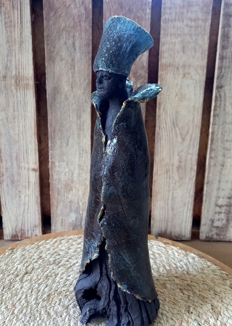 May include: A ceramic sculpture of a person wearing a dark blue robe with gold accents and a teal hat. The sculpture is standing on a wooden base.