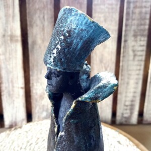 May include: A ceramic sculpture of a person wearing a blue and black hat and a blue and black robe. The sculpture has a textured surface and is painted in shades of blue and black.