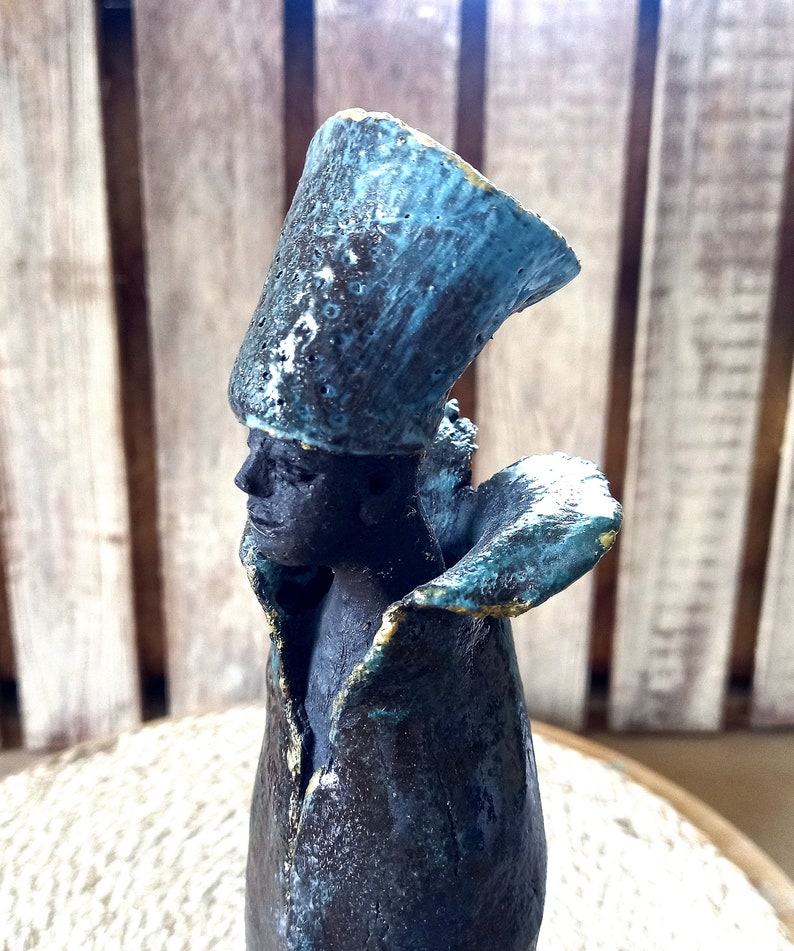 May include: A ceramic sculpture of a person wearing a blue and black hat and a blue and black robe. The sculpture has a textured surface and is painted in shades of blue and black.