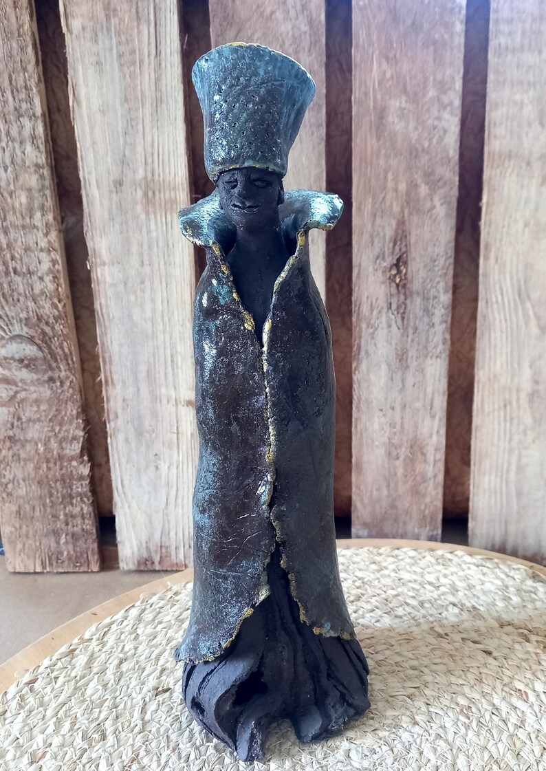 May include: A ceramic sculpture of a person wearing a tall, blue hat and a long, dark robe with gold accents. The sculpture has a textured surface and is standing on a wooden base.