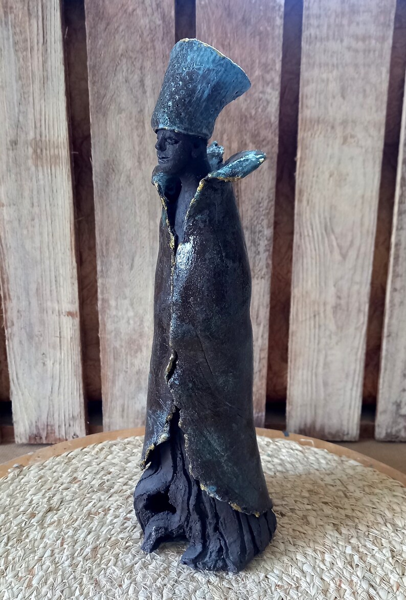May include: A ceramic sculpture of a person wearing a blue and brown robe with a tall, blue hat. The sculpture has a textured surface and is standing on a woven mat.