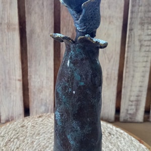 May include: A ceramic vase with a unique, abstract design. The vase is a dark brown colour with a teal glaze and a textured surface. The top of the vase is shaped like a flower.