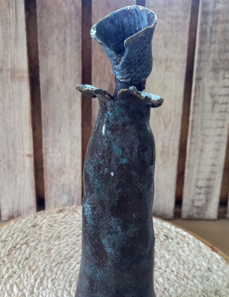 May include: A ceramic vase with a unique, abstract design. The vase is a dark brown colour with a teal glaze and a textured surface. The top of the vase is shaped like a flower.