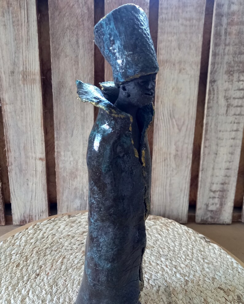 May include: A ceramic sculpture of a person wearing a tall, blue hat and a long, dark blue robe with gold accents. The sculpture has a textured, aged look.