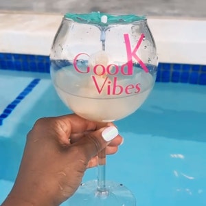 May include: A clear glass with a teal lid and a pink sticker that says "Good Vibes" is held by a hand in a swimming pool.