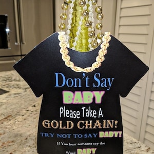 May include: A black baby onesie shaped sign with the text "Don't Say Baby Please Take A Gold Chain! Try Not To Say Baby! If You Hear Someone Say The Word Baby Take Their Gold Chain. The Person With The Most Gold Chains At The End Of The Shower Wins!"