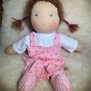 Könnte beinhalten: Eine handgemachte Puppe mit braunen Haaren, blauen Augen und rosigen Wangen. Die Puppe trägt ein weißes Hemd und einen rosa Overall mit einem weißen Kreuzmuster. Die Beine der Puppe sind mit rosa gehäkelten Socken bedeckt.