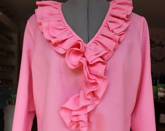 May include: A pink blouse with a large ruffled collar. The collar is made of multiple layers of fabric and is gathered at the center.