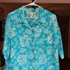 May include: A turquoise button-down shirt with a white floral print. The shirt has a pocket on the left side and a label that reads "MIRROR, MIR STUDIO" and "SIZE M (LAD)".