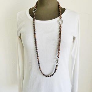 May include: A long necklace featuring wooden beads and brown chain links. The necklace has silver-coloured metal accents and is displayed on a white top.