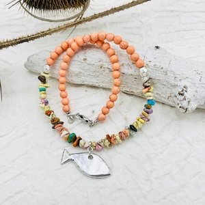 May include: A peach-coloured beaded necklace with a silver fish charm. The necklace is made with a variety of colourful stones, including turquoise, pink, and brown.