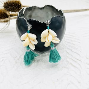 May include: A pair of earrings with a cluster of white seashells and a teal tassel hanging from each. The earrings are silver and have a small green bead at the top.