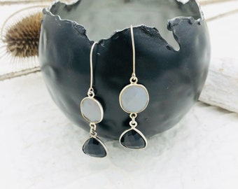 May include: Silver-toned dangle earrings featuring a light gray, circular stone above a black, triangular stone. The earrings are displayed against a dark gray, textured bowl.