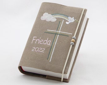 May include: A brown linen book cover with a white cross, a rainbow, and a dove embroidered on it. The cover also has the name "Frieda" and the year "2022" embroidered on it.