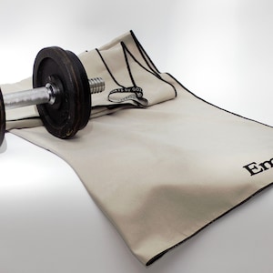 Könnte beinhalten: Eine Hantel mit schwarzen Gewichtsscheiben liegt auf einem beigen Trainingstuch. Das Handtuch hat einen schwarzen Rand und den Namen "Emma" in Schwarz gestickt. Der Text "OATS BY GO2" ist ebenfalls vorhanden.