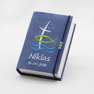 Könnte beinhalten: Ein blauer Stoffbuchumschlag mit einem weißen Kreuz, einem grünen Fisch und blauen Wellen, die darauf gestickt sind. Der Name "Niklas" und das Datum "15.04.2018" sind ebenfalls auf dem Umschlag gestickt.
