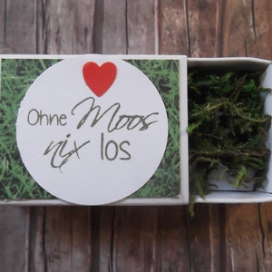 Könnte beinhalten: Eine weiße Streichholzschachtel mit grünem und weißem Muster. Der Deckel hat einen weißen Kreis mit einem roten Herzen und dem Text "Ohne Moos nix los". Die Schachtel ist geöffnet und enthält grünes Moos.