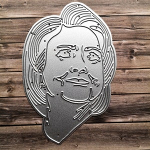 Puede incluir: Un troquel de metal plateado de la cara de una mujer con un contorno detallado de su cabello, ojos, nariz y boca.