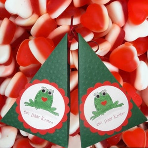 Könnte beinhalten: Zwei grüne dreieckige Geschenkboxen mit gewellten Kanten. Jede Box hat einen weißen Kreis mit einem Comic-Frosch, der ein Herz hält, und den Text "ein paar Kroten". Die Boxen sind von roten und weißen herzförmigen Süßigkeiten umgeben.