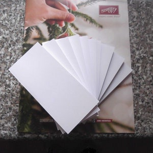 May include: A stack of ten white cardstock sheets, each measuring approximately 4.25 inches by 5.5 inches. The cardstock is arranged in a fan shape, with the top sheet slightly overlapping the sheet below it. The cardstock is on top of a magazine with the text "Stampin' Up!" printed on the cover.