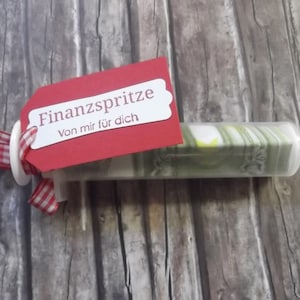 Può includere: Un nastro gingham rosso e bianco legato attorno a una siringa di plastica trasparente piena di contanti. Un'etichetta rossa con lettere bianche recita "Finanzspritze Von mir für dich".