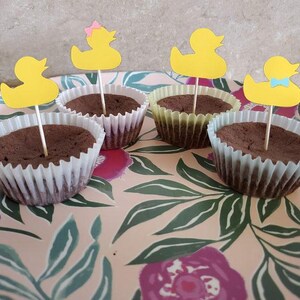 May include: Four chocolate cupcakes in white and yellow paper liners, each topped with a yellow duck-shaped pick. The cupcakes sit on a pink floral patterned tray, creating a festive and playful presentation.