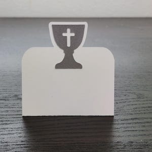 May include: A white card holder with a gray chalice and cross design. The chalice is at the top of the card, and the cross is in the center of the chalice. The card holder is on a dark wooden surface.
