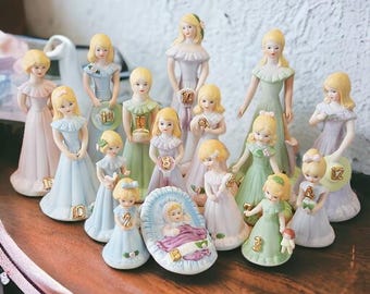 Op de afbeelding: Een verzameling van 15 porseleinen beeldjes van meisjes gekleed in pastelkleurige jurken. Elk beeldje houdt een nummer vast, variërend van 1 tot 15. De beeldjes zijn in een cirkel op een houten oppervlak gerangschikt.